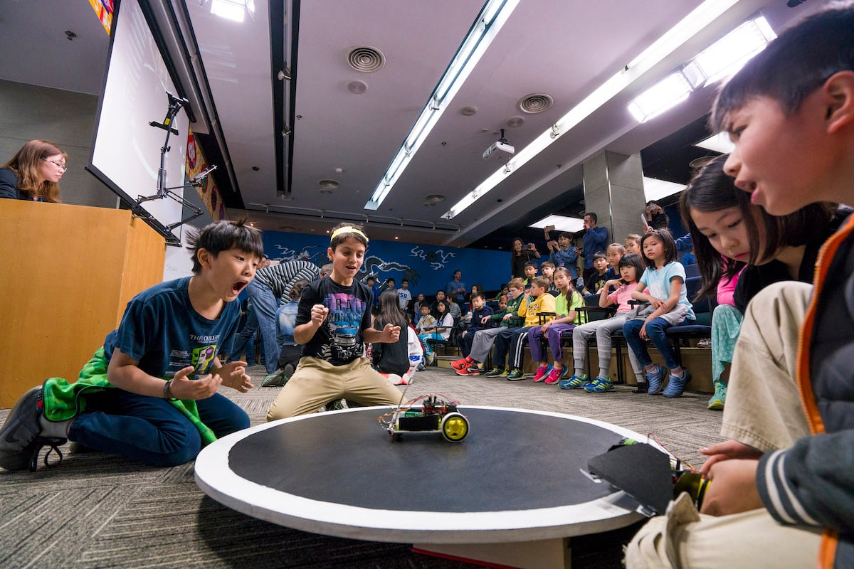 Students play excitedly with robots in the foreground while others watch in the background